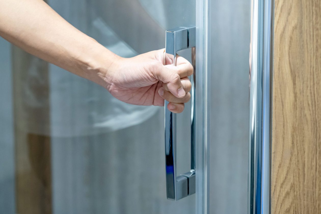 Customer hand holding chrome door handle opening transparent glass door of modern shower room in home design store. Sanitary ware for bathroom interior design